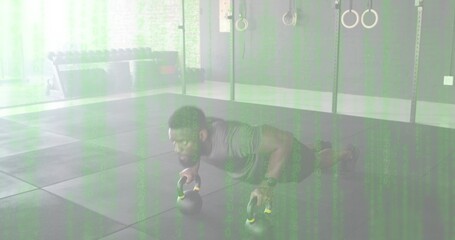 Performing man stabilizing kettlebell push-up in gym on rubber tiles, wearing gray shirt and watch