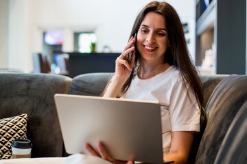 Female smiling woman working on a laptop while talking on the phone, showing multitasking, remote work, digital communication and modern professional lifestyle in a cozy indoor workspace setting.
