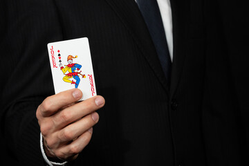 Close up to a businessman on suit holding a joker card on his hand at the left side of the screen