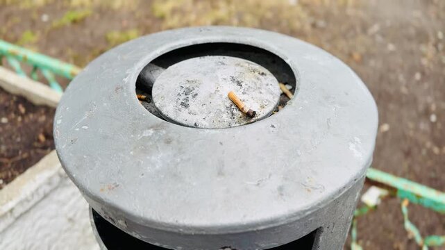 Cigarette butt on metal ashtray atop public trash can, ash residue and crushed tip visible, green railing and soil in background, dull overcast light, damp pavement, neglected city