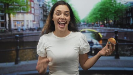 Woman waves hand while holding muffin on a canal street in amsterdam, smiling and beckoning to come closer; joyful invitation.