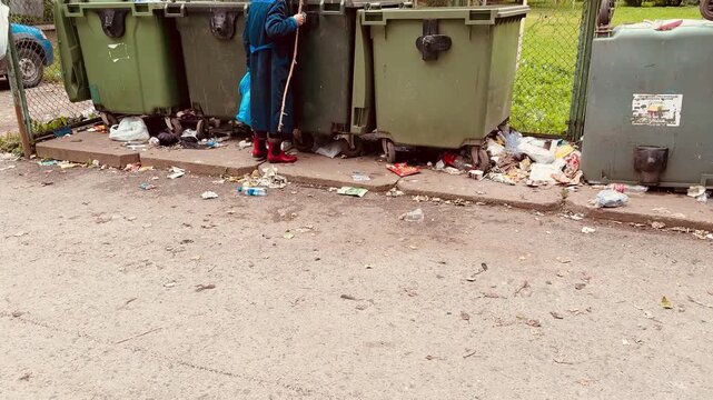 Elderly white woman rummaging dumpster in urban alley, worn blue coat and red boots, cane for support, blue plastic bag, scattered food wrappers and bottles, overflowing green