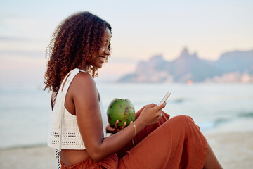 Woman relaxing on a beach lounge with fresh coconut water and using a smartphone during sunset in Rio de Janeiro, Brazil