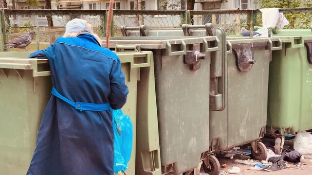 Elderly white woman rummaging dumpster in urban alley, worn blue coat and red boots, cane for support, blue plastic bag, scattered food wrappers and bottles, overflowing green