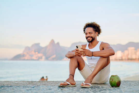 Young man relaxing on Ipanema beach, feeling happy while using his phone with a fresh coconut by his side