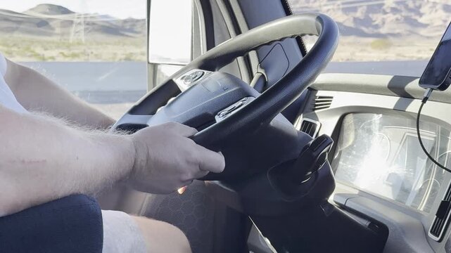 Male truck driver's hands on the steering wheel while driving. Scenic mountains and oncoming traffic are visible through the window. Professional logistics and transportation atmosphere in motion.