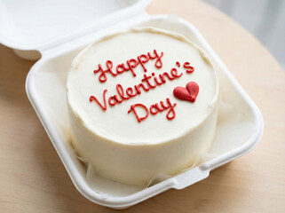 A small white cake with red 'Happy Valentine's Day' frosting and a heart, presented in an open white takeout container.