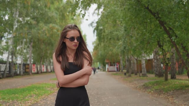 white woman in black dress standing on treelined park path, arms crossed and sunglasses, projecting aloof attitude later encounters white teenage male in hoodie, delivers pointed finger touch