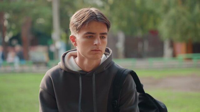 white teen student sitting bench in park, wearing dark hoodie and backpack, pensive expression, late afternoon sunlight, shallow depth of field, quiet playground in background, cinematic portrait