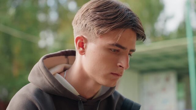 teen boy in hoodie looking down, cinematic park portrait with pensive expression, soft golden light and shallow depth of field, student waiting at bus stop, candid closeup capturing vulnerability