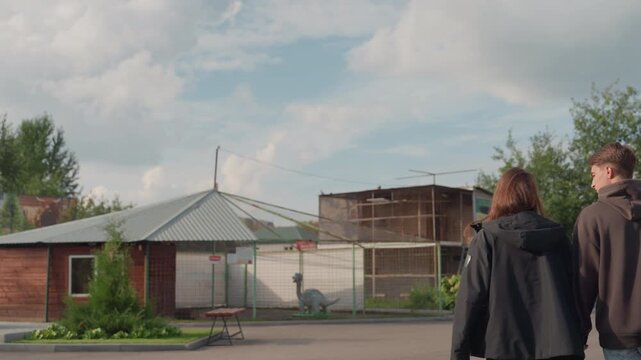 two white teenagers walk past pavilion under cloudy summer sky, chainlink fence and rusted booths frame scene, string lights droop above, backpacks and hoodies, slow reflective stroll, suburban