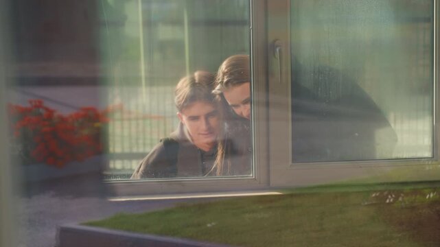 white teens peering through window, soft morning light and reflective glass reveal closeup faces, garden planter outside, hushed curiosity and quiet bond between siblings, cinematic composition