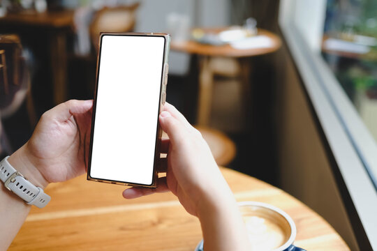 A person holds a large smartphone with a blank white screen near a cafe window, with a cup of coffee on the table.