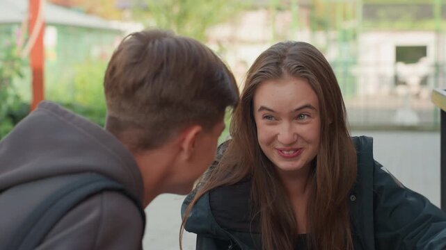 caucasian teens at exhibit window sharing secret, boy and girl exchange mischievous smiles and whispers, student hangout vibe with jackets and backpacks, candid conversation shifts to playful peek