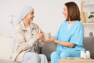 Obraz premium Young woman after chemotherapy with cup of tea and nurse on sofa at home. Brain cancer concept