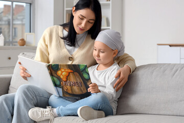 Naklejka premium Little girl after chemotherapy with her mother reading book on sofa at home. Childhood cancer awareness concept