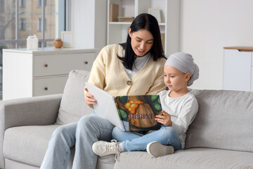 Naklejka premium Little girl after chemotherapy with her mother reading book on sofa at home. Childhood cancer awareness concept