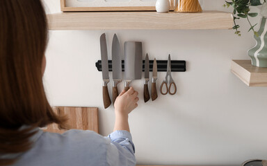 Young woman taking knife from magnetic holder on light wall in kitchen