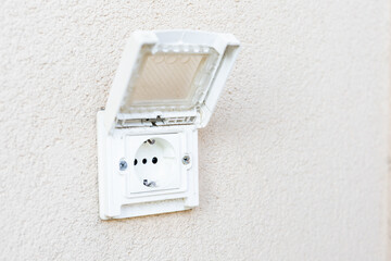 A white outdoor electrical power socket with an open protective plastic cover is installed on a textured beige stucco wall and photographed in bright natural light