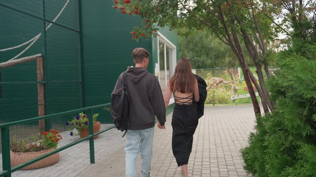 white couple holding hands garden path, young white man in hoodie with backpack and woman in black backless dress walking toward green enclosure, potted planters and brick pavers, casual campus date