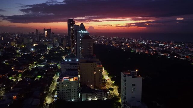 Cinematic aerial drone view of the Santo Domingo skyline at sunrise. The city lights glow against the dramatic sky as dawn breaks over the ocean, Dominican Republic