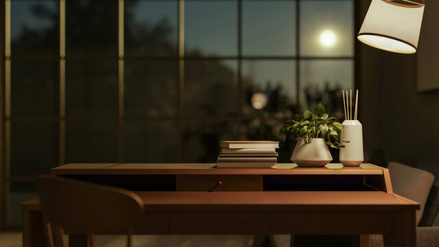 Books and pot plant with diffuser on wooden table under lamp in dim room and sky through glass wall.
