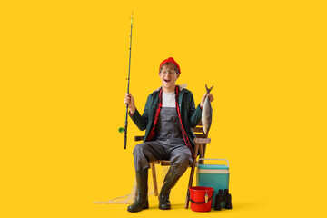 Funny young man with fishing rod and caught fish sitting in chair on yellow background