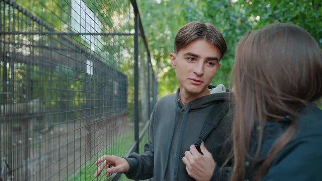 young caucasian man chats by fence in park, casual hoodie and backpack, gesturing and smiling at female friend, warm greenery backdrop, easygoing banter and laughter, spontaneous friendly connection