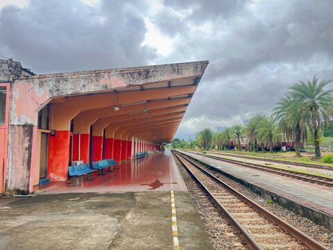 A perspective view of a long, empty railway platform featuring orange pillars and blue seats. Parallel tracks stretch toward palm trees under a dramatic, overcast tropical sky