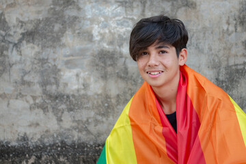 A young person wrapped in a rainbow flag, symbolizing LGBTQ+ pride, stands against a textured wall....