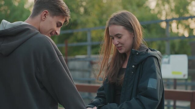 two teens exchanging item by railing in evening, casual jackets and shy smiles create tender atmosphere candid closeups capture gentle hand gestures, soft golden backlight, parklike surroundings