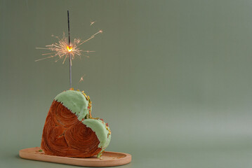Festive holiday candle sparkler birthday anniversary celebration Heart shaped pistachio croissant on green background with copy space. Puff pastry new trendy sweet dessert bakery. Creamy dough