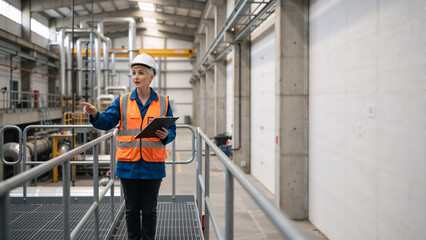 Female industrial engineer in safety vest and hard hat pointing at machinery. Professional woman inspector with clipboard in a factory. Facility management and safety inspection concept
