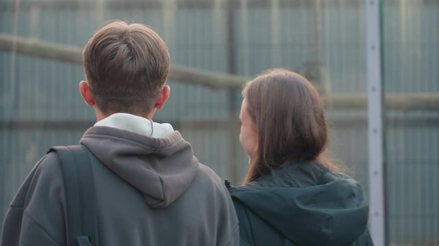 two white teens read park sign checking map as students companion smiles while pointing at enclosure backpacks and hoodies visible evening urban zoo setting with metal fence and glass panels