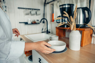 woman hands places a white bowl on a wooden table in a cozy kitchen. A vase with dried wheat and a coffee maker are visible.