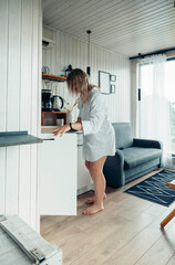 young Caucasian woman with long brown hair stands in a cozy vacation home, reaching for items in a white cabinet. Natural light fills the room, creating a warm atmosphere