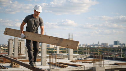 Construction worker walking on a steel beam carrying a wooden plank. Professional builder in a hard hat at a high-rise building site. Urban development and manual labor concept