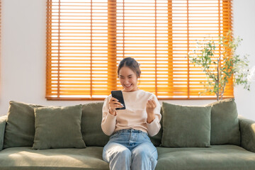 Happy young asian woman relax on comfortable couch at home texting messaging on smartphone, smiling girl use cellphone, chatting online message, shopping online from home