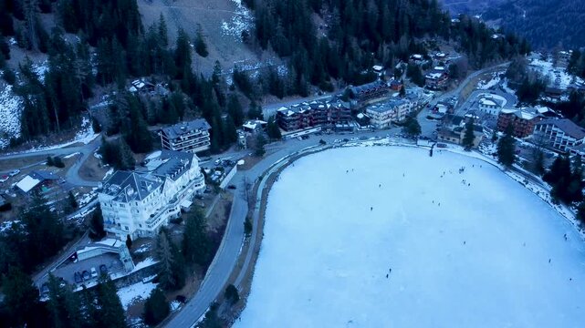 Aerial drone footage of a frozen lake in Champex, Switzerland, during winter. People ice skating surrounded by snowy alpine scenery. Shot in 4K using a DJI Mini 2.