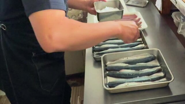 A chef sprinkling salt over fresh fish fillets arranged in metal trays