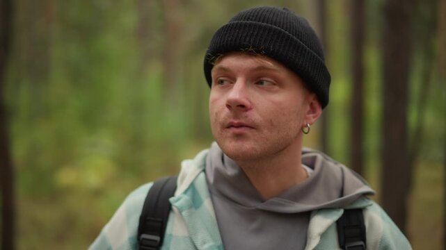 frontal portrait of man speaking in forest with expressive gaze and slight smile, wearing hoodie and beanie with backpack straps visible, vlogger tone and natural ambient atmosphere among pines.