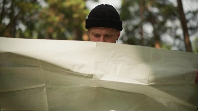 Hiker reading paper map in forest solo traveler wearing knit cap examines large folded map, scanning routes among pine trees, sunlight filtering through canopy, thoughtful expression, plotting route,