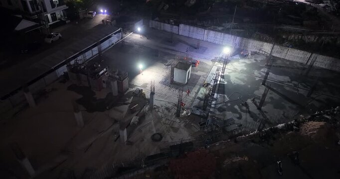 Aerial night view of workers on a large building foundation illuminated by floodlights.
