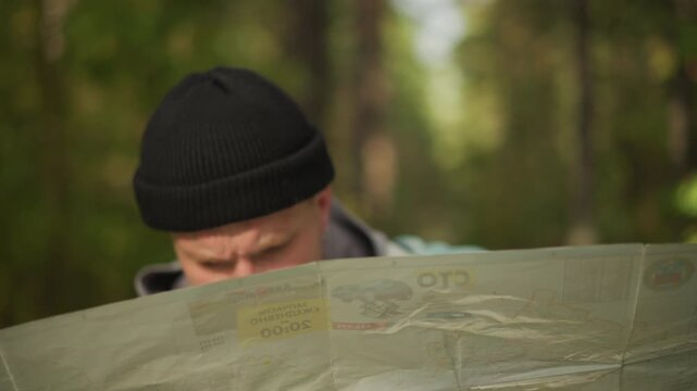 white hiker studies folded map intently in shaded forest, closeup beanie portrait and hooded jacket, concentrating on coordinates and contours, weighing options and selecting next waypoint,