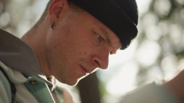 Caucasian hiker studying map outdoors, sunlit forest background, beanie and backpack, focused expression and stubble, earring detail, morning light, campsite mood, quiet contemplation and route