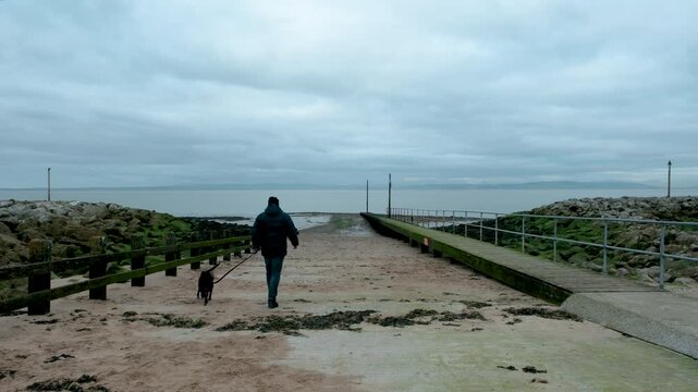 Man walking his lurcher dog away from the beach in Morecambe on a grey overcast winter day in December