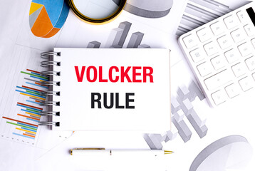 Volcker rule phrase on notepad surrounded by charts, magnifying glass, pen and white calculator