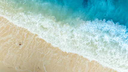 Aerial video shows waves crashing against a tropical coastline with turquoise, clear waters contrasting with a bright sky, and coral reefs on the shores of a tropical island with emerald waters.	