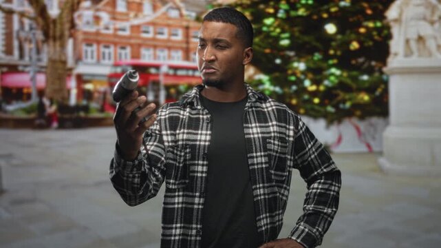 Man holding electric screwdriver with right hand, wearing plaid shirt, studying drill tip while hand rests on hip on street near a decorated christmas tree; concentration repair.