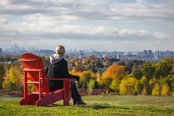 Obraz premium Middle-aged woman sitting on a red wooden chair atop a hill, enjoying a panoramic autumn view of Toronto’s skyline and colorful trees
